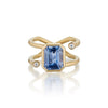 A gold ring featuring a large rectangular blue sapphire with lavender undertones center stone and two small round clear accent stones on the band, shown on a clean white background with a soft reflection.