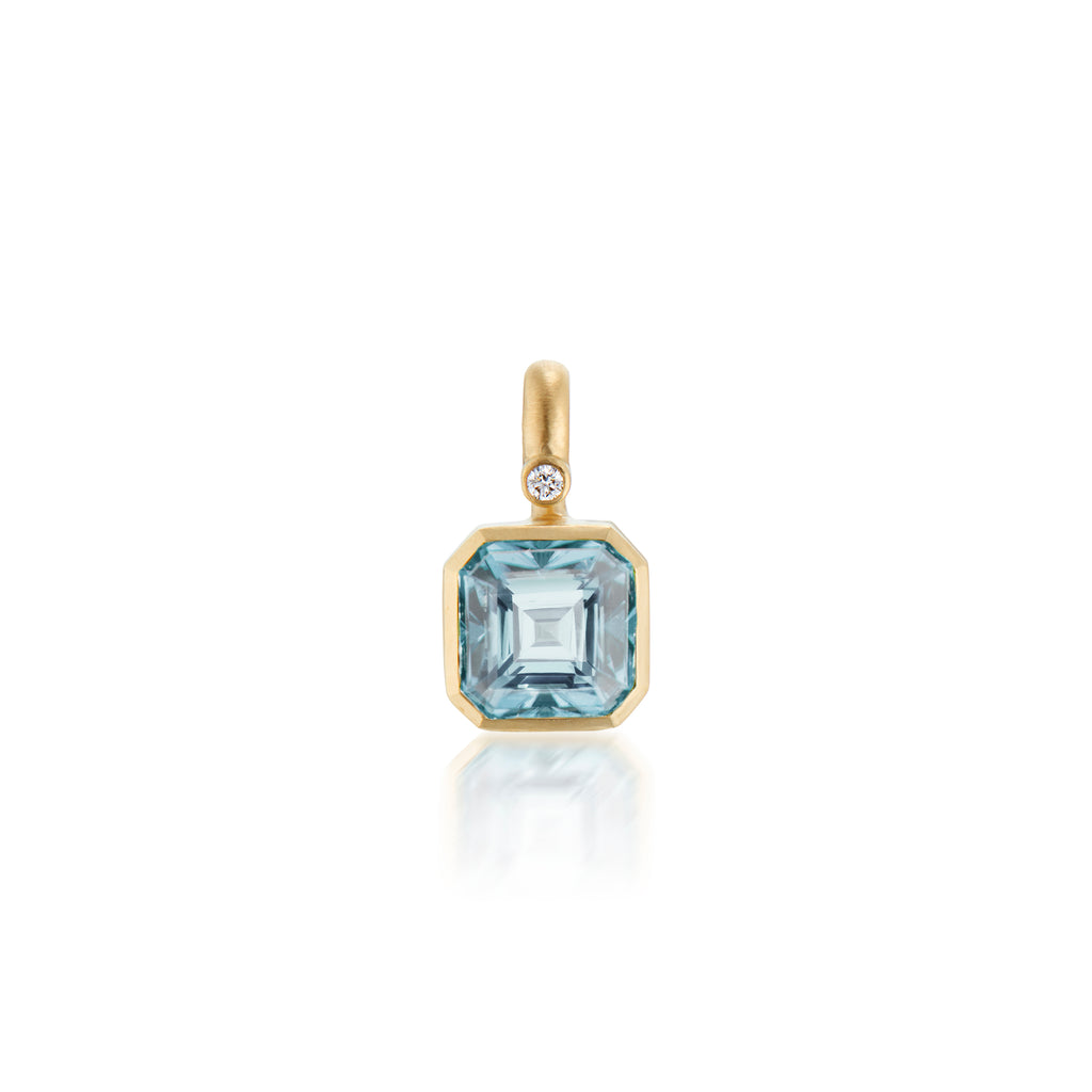 A gold pendant featuring a square-cut blue aquamarine gemstone and a small round diamond accent, shown on a clean white background with a soft reflection below.