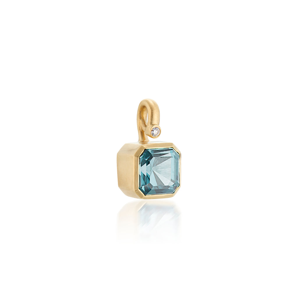 A gold pendant featuring a faceted light blue aquamarine gemstone in a bezel setting with a small round accent stone, shown on a clean white background with a soft reflection.
