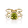 A gold ring featuring a large, faceted emerald-cut olive green Peridot or blue aquamarine center stone, with two wraparound gold bands adorned with small round-cut diamonds on either side, set against a clean white background.