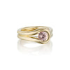 A gold ring with a unique, flowing double-band design and a round pink Malaya Garnet gemstone, shown on a clean white background with a soft reflection.