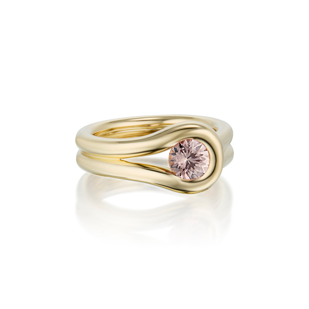 A gold ring with a unique, flowing double-band design and a round pink Malaya Garnet gemstone, shown on a clean white background with a soft reflection.