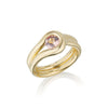 A gold ring with a unique, flowing double-band design and a round pink Malaya Garnet gemstone, shown on a clean white background with a soft reflection.