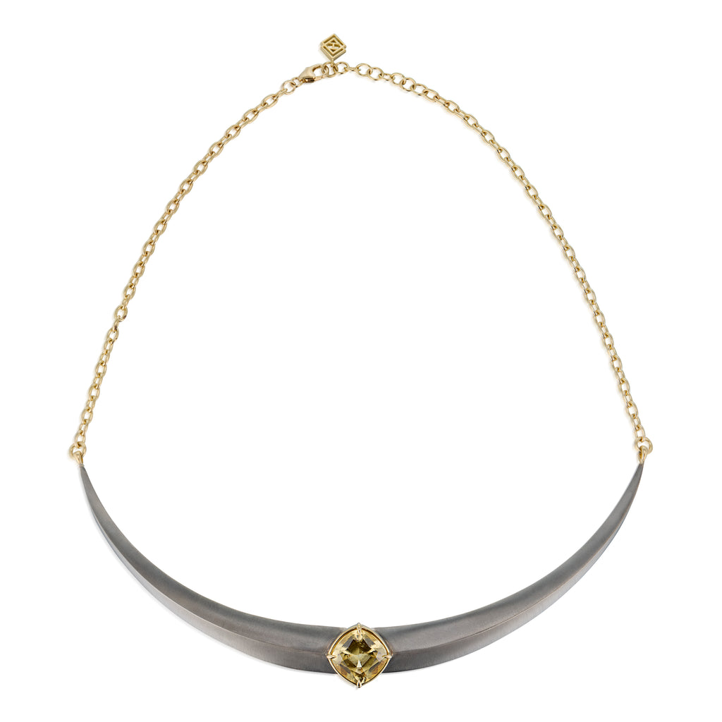 A curved collar necklace featuring a matte gray horn-like center pendant with a central gold gemstone, grossular garnet, suspended from a delicate gold chain against a white background.
