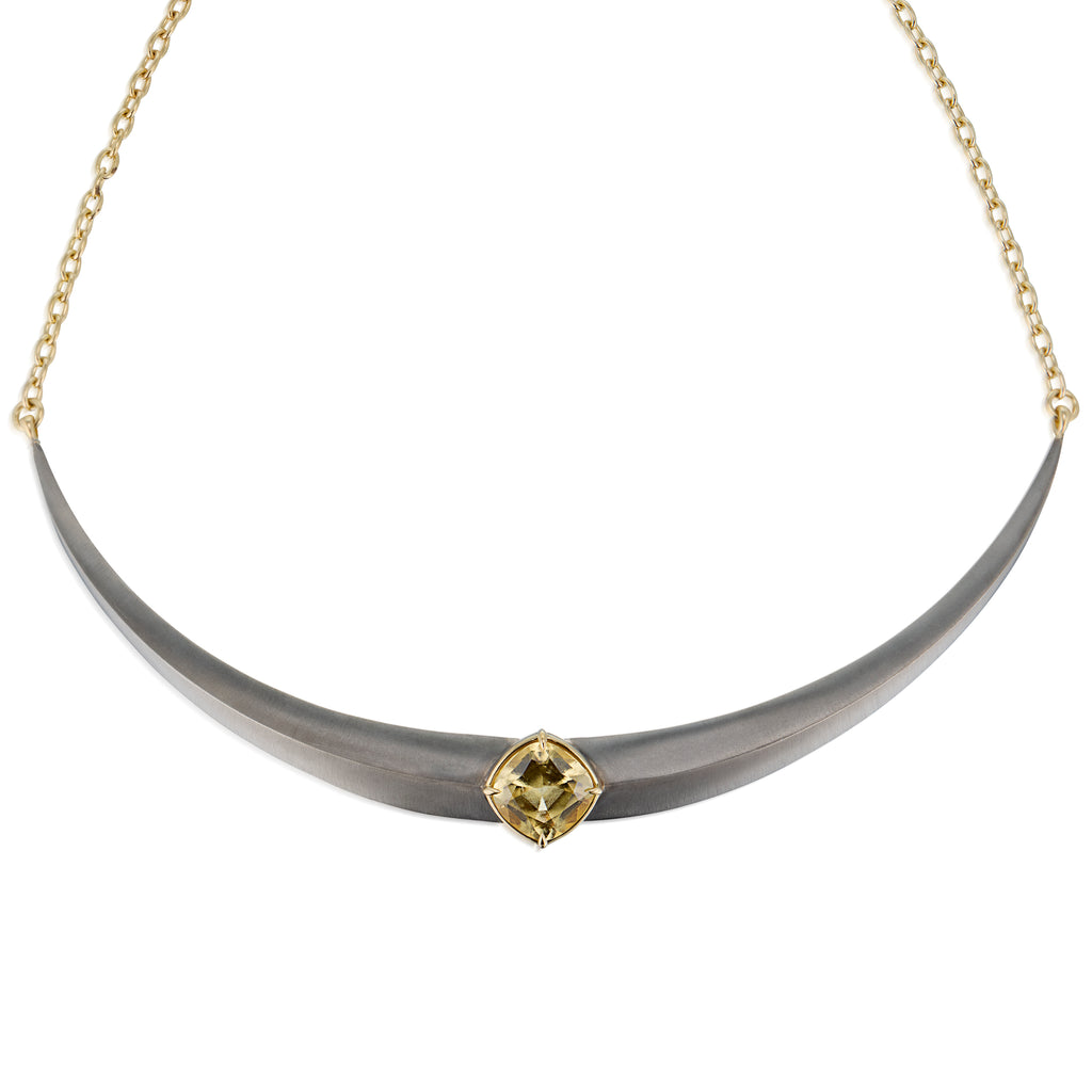 A curved collar necklace featuring a matte gray horn-like center pendant with a central gold gemstone, grossular garnet, suspended from a delicate gold chain against a white background.