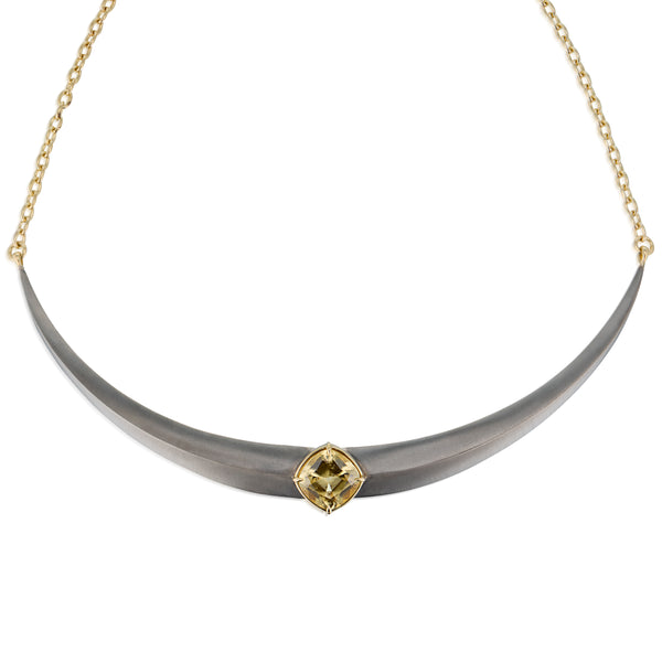 A curved collar necklace featuring a matte gray horn-like center pendant with a central gold gemstone, grossular garnet, suspended from a delicate gold chain against a white background.